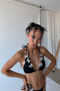 Woman wearing a black bikini with white floral details in a minimal indoor setting.