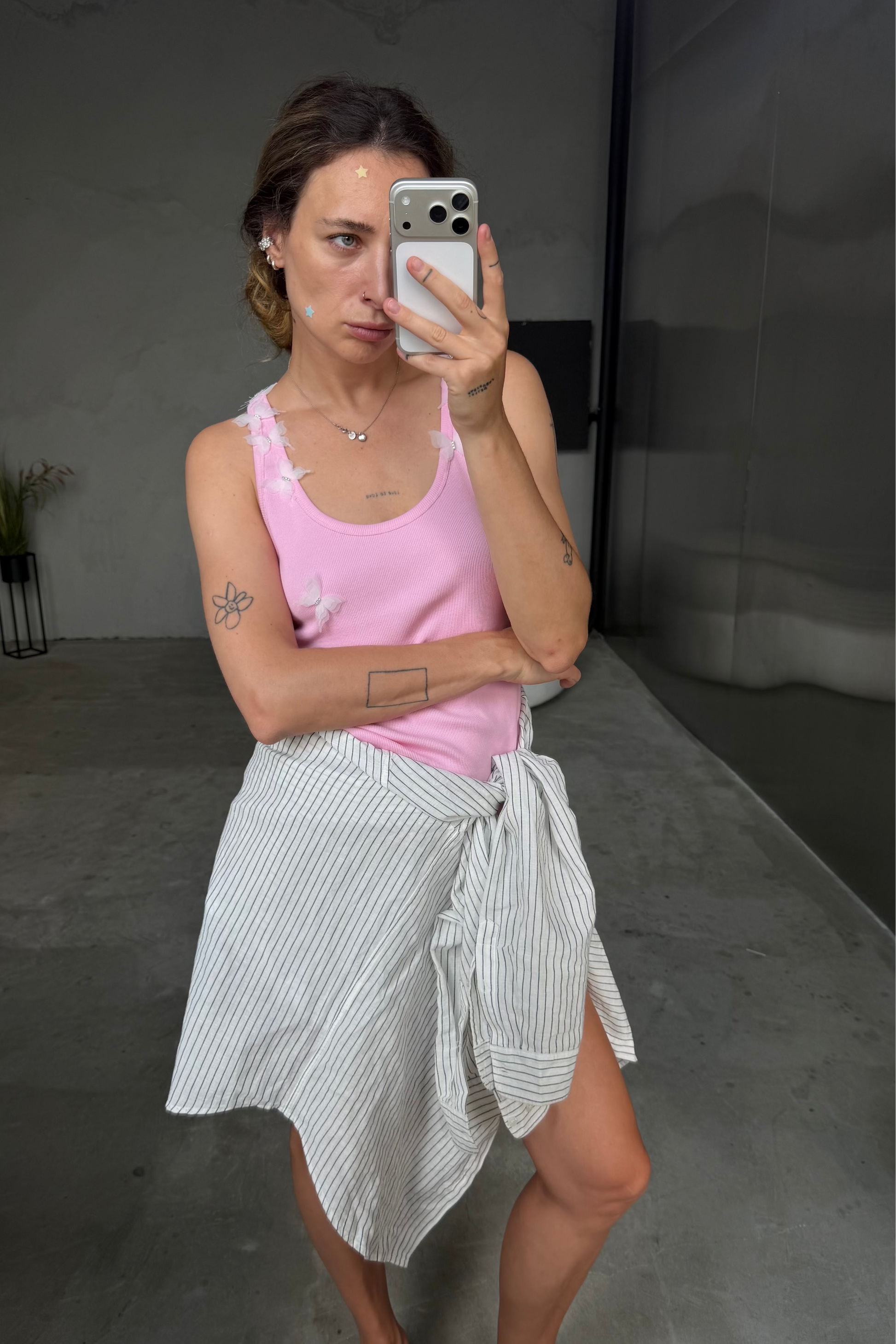 A woman takes a mirror selfie wearing a light pink ribbed tank top and a white shirt with thin black vertical stripes tied around her waist as a skirt. She is wearing black leather mule flats in a minimalist gray room.