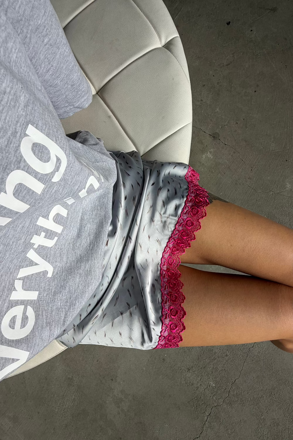 A person reclining on a white tufted chair wearing a gray satin pajama skirt with a bright pink lace trim, paired with a gray graphic t-shirt.