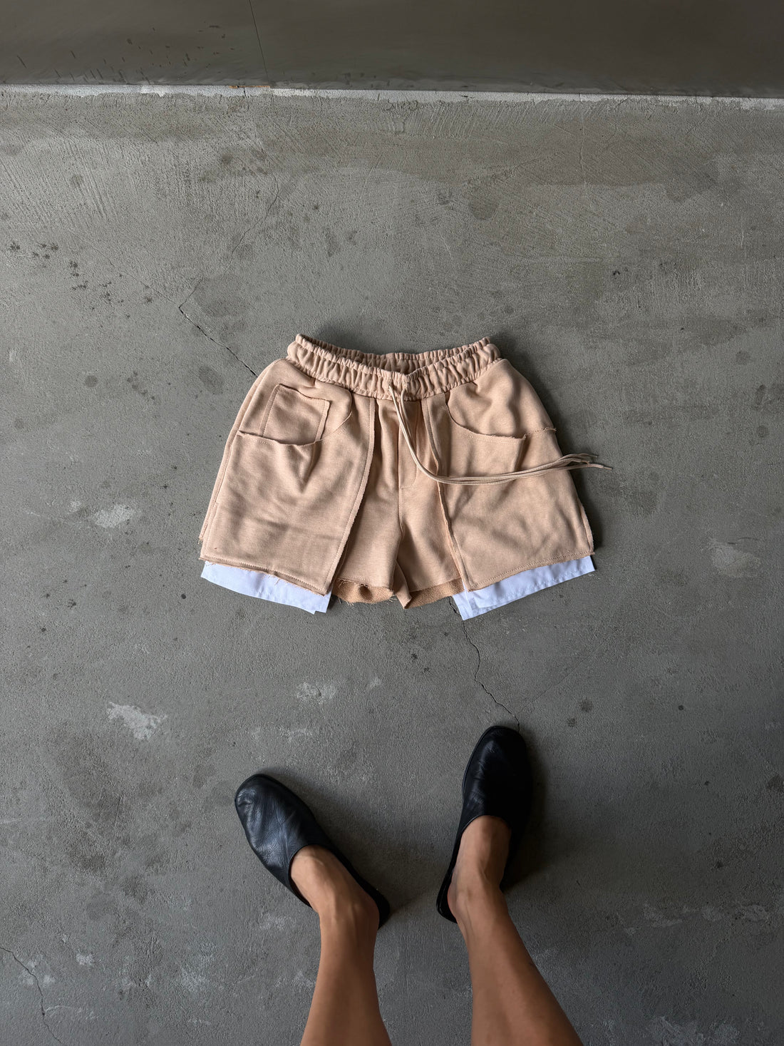 A pair of beige drawstring shorts with a relaxed fit, laid flat on a textured gray concrete surface. The shorts feature an elastic waistband, side pockets, and a white inner layer peaking out at the hem.