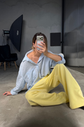 A woman sitting on a concrete floor in a studio setting, wearing a light blue oversized button-up shirt and bright yellow wide-leg pants, holding a phone to take a mirror selfie.