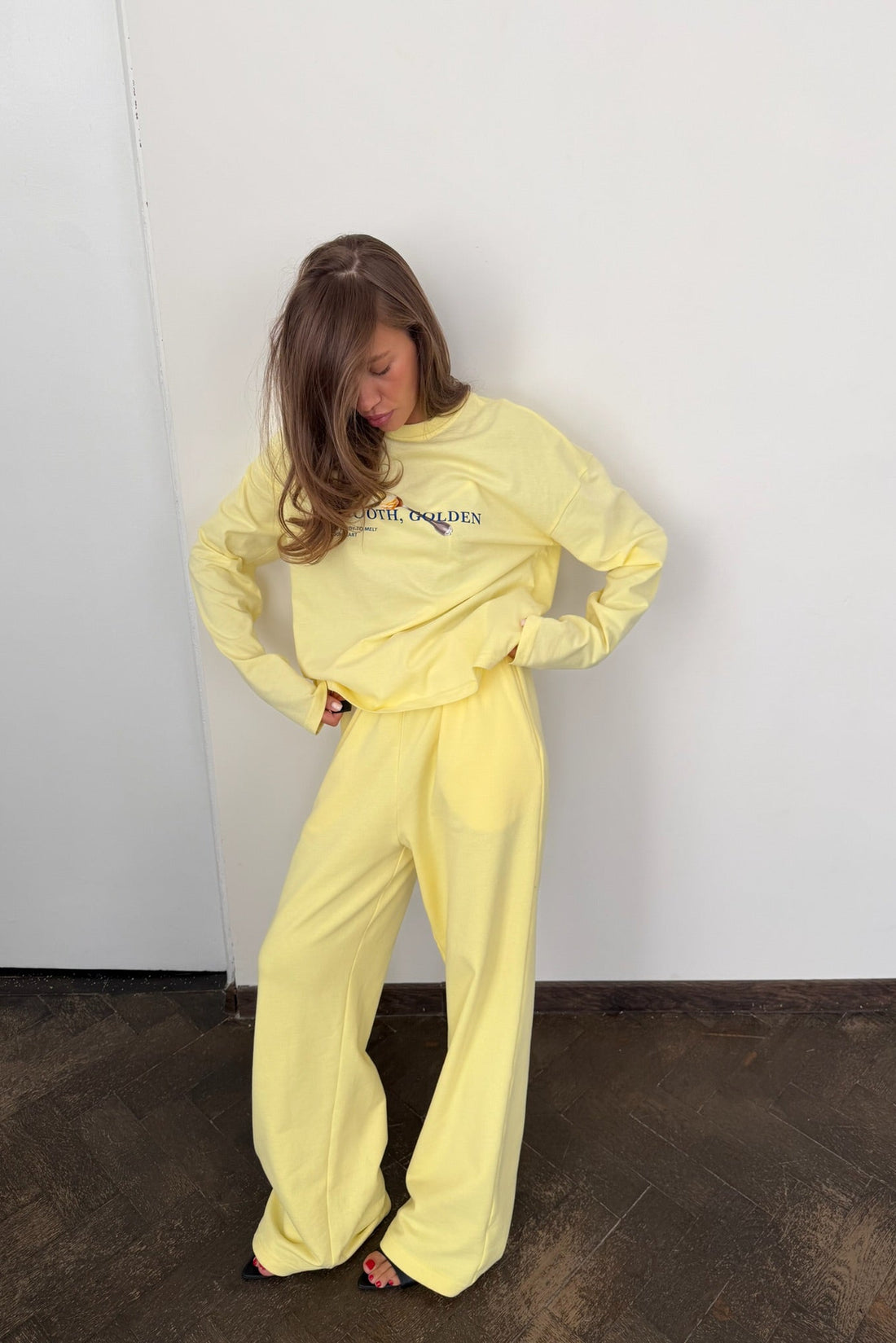 A person wearing a pale yellow long-sleeve sweatshirt and matching sweatpants. The sweatshirt has a graphic on the chest with the text 'SMOOTH, GOLDEN' and a small illustration.