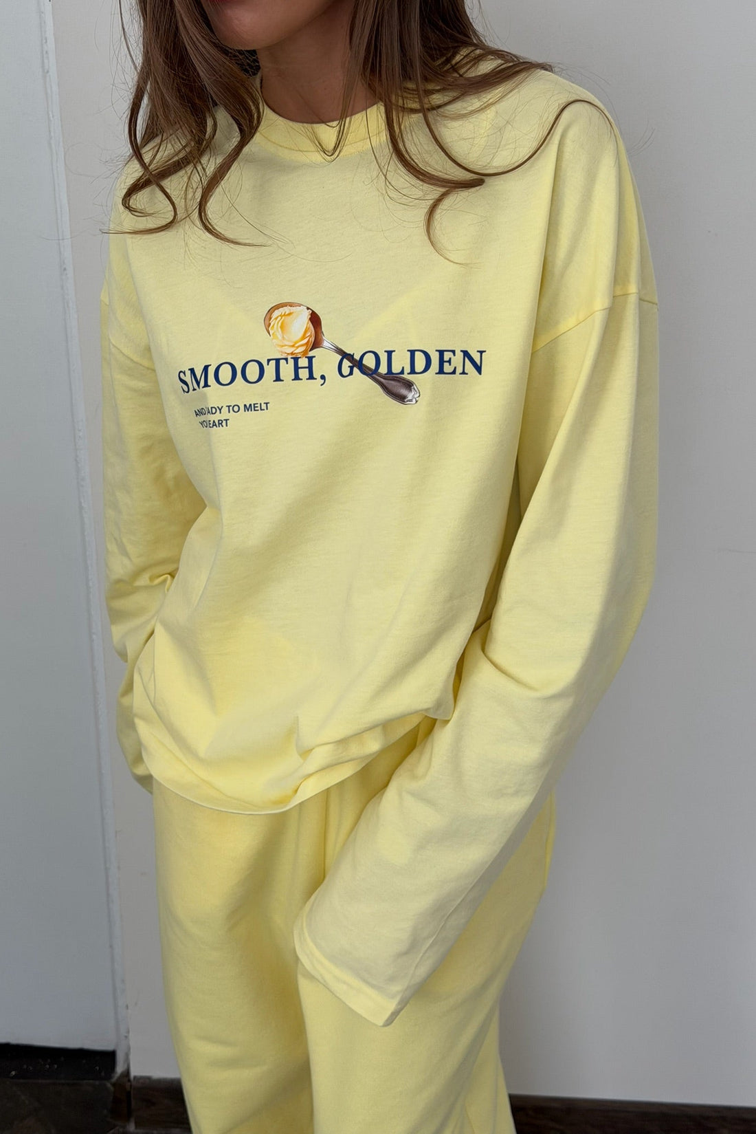 A person wearing a pale yellow long-sleeve sweatshirt and matching sweatpants. The sweatshirt has a graphic on the chest with the text 'SMOOTH, GOLDEN' and a small illustration.