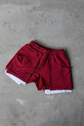 A pair of deep red drawstring shorts with white inner lining or underlayer peeking out at the hem, laid flat on a textured gray concrete surface.