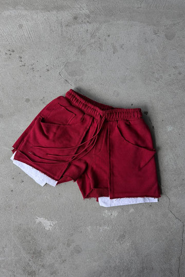 A pair of deep red drawstring shorts with white inner lining or underlayer peeking out at the hem, laid flat on a textured gray concrete surface.