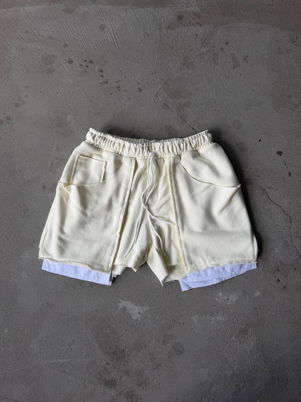 Cream-colored drawstring shorts with a raw, frayed hem and a light blue undershirt peeking out at the bottom, laid flat on a textured gray concrete surface.