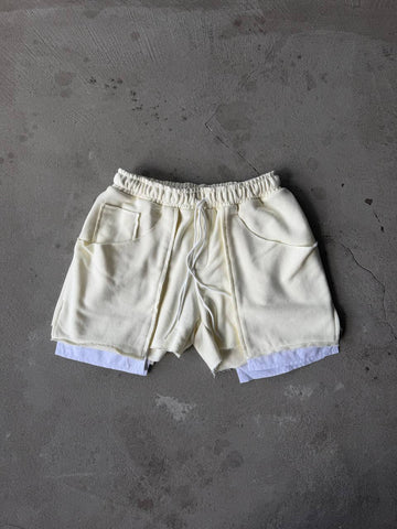 Cream-colored drawstring shorts with a raw, frayed hem and a light blue undershirt peeking out at the bottom, laid flat on a textured gray concrete surface.