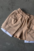 A pair of beige drawstring shorts with a relaxed fit, laid flat on a textured gray concrete surface. The shorts feature an elastic waistband, side pockets, and a white inner layer peaking out at the hem.