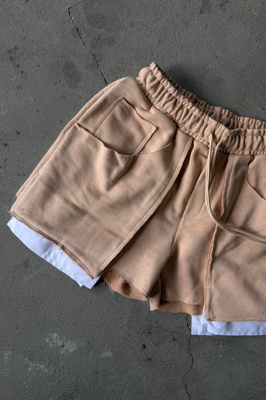A pair of beige drawstring shorts with a relaxed fit, laid flat on a textured gray concrete surface. The shorts feature an elastic waistband, side pockets, and a white inner layer peaking out at the hem.